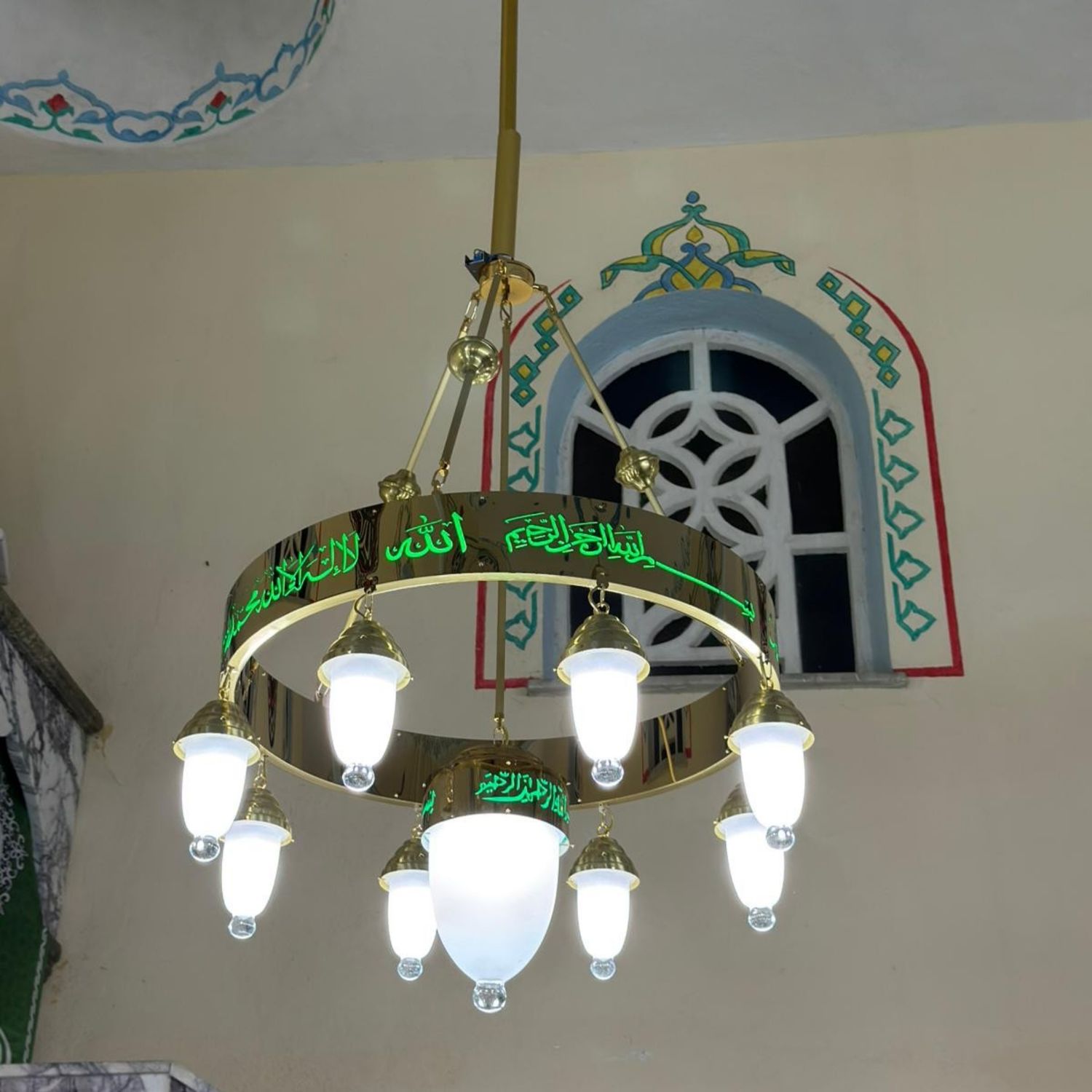 Tek halkalı 80 cm yeşil tevhidli ledli cami avizesi cami içinde, kalem işli duvarın önünde yanarken