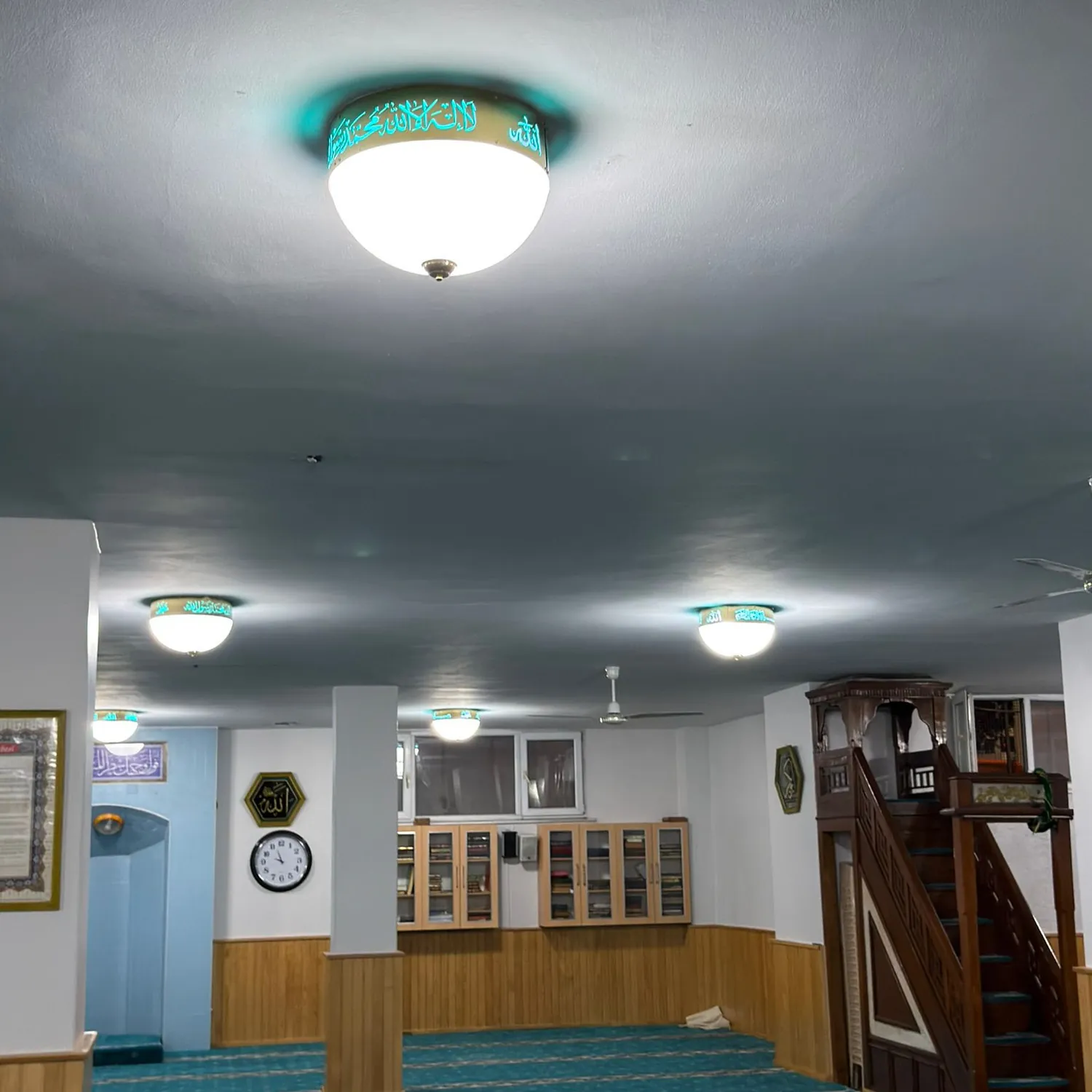 Turkuaz halılı cami iç mekânında minber ve kitaplığın üstünde sıralanmış turkuaz Kelime-i Tevhidli plafonyerler